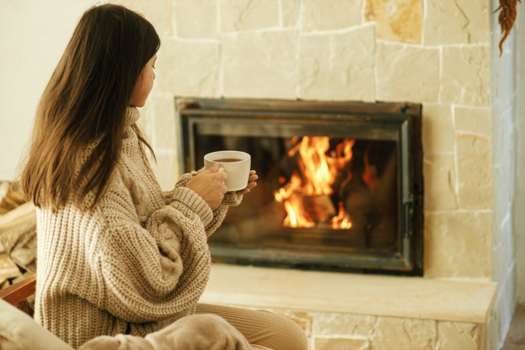 Woman in cozy sweater holding cup of warm tea at fireplace in rustic room. Heating house in winter with wood burning stove. Young stylish female relaxing at fireplace in farmhouse